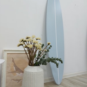 K&ouml;nnte beinhalten: Ein hellblaues Surfbrett mit einem vertikalen wei&szlig;en Mittelstreifen, das an einer Wand lehnt. Im Vordergrund steht eine wei&szlig;e, strukturierte Vase mit einem Strau&szlig; aus getrockneten cremefarbenen Blumen und gr&uuml;nen Eukalyptuszweigen neben einem Walbild.