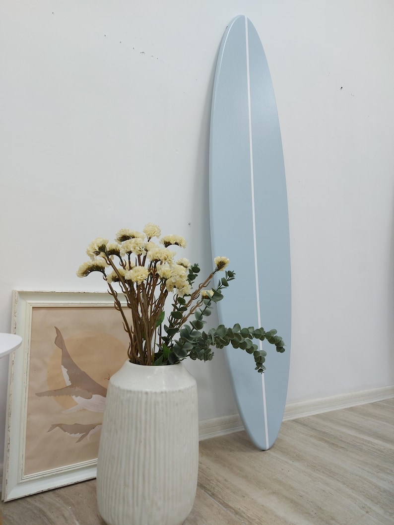 K&ouml;nnte beinhalten: Ein hellblaues Surfbrett mit einem vertikalen wei&szlig;en Mittelstreifen, das an einer Wand lehnt. Im Vordergrund steht eine wei&szlig;e, strukturierte Vase mit einem Strau&szlig; aus getrockneten cremefarbenen Blumen und gr&uuml;nen Eukalyptuszweigen neben einem Walbild.