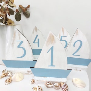 May include: Five wooden sailboat table numbers, painted white with blue accents. The numbers 1, 2, 3, 4, and 5 are painted in blue on the sail of each sailboat.