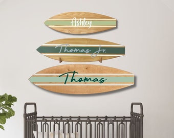 May include: Three wooden surfboard-shaped wall decorations featuring horizontal stripes in shades of green and white. The top board displays the name Ashley, the middle board displays Thomas Jr., and the bottom board displays the name Thomas in script.