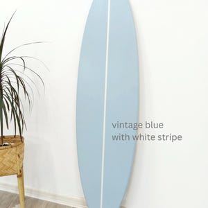 K&ouml;nnte beinhalten: Ein Surfbrett mit einer glatten, hellblauen Oberfl&auml;che und einem einzelnen vertikalen wei&szlig;en Streifen in der Mitte. Das Brett zeigt den Text vintage blue with white stripe, der in einer einfachen grauen Schrift auf der rechten Seite steht.