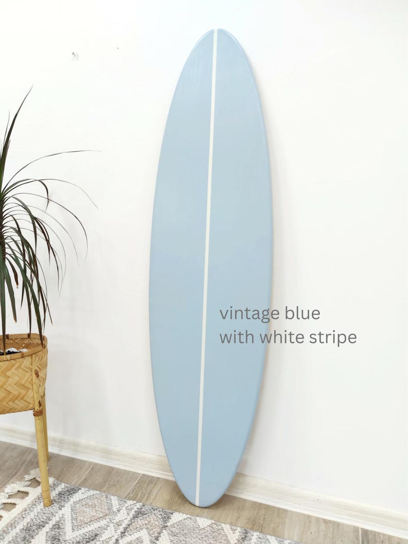 K&ouml;nnte beinhalten: Ein Surfbrett mit einer glatten, hellblauen Oberfl&auml;che und einem einzelnen vertikalen wei&szlig;en Streifen in der Mitte. Das Brett zeigt den Text vintage blue with white stripe, der in einer einfachen grauen Schrift auf der rechten Seite steht.