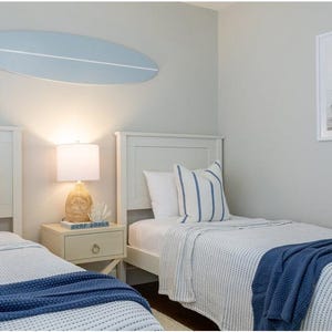 K&ouml;nnte beinhalten: Ein Schlafzimmer mit zwei wei&szlig;en Betten, blau-wei&szlig; gestreiften Kissen und marineblauen &Uuml;berw&uuml;rfen. &Uuml;ber den Betten h&auml;ngt eine hellblaue Surfbrett-Wanddekoration. Zwischen den Betten steht ein Nachttisch mit Lampe, an der Wand h&auml;ngt ein gerahmtes Bild.