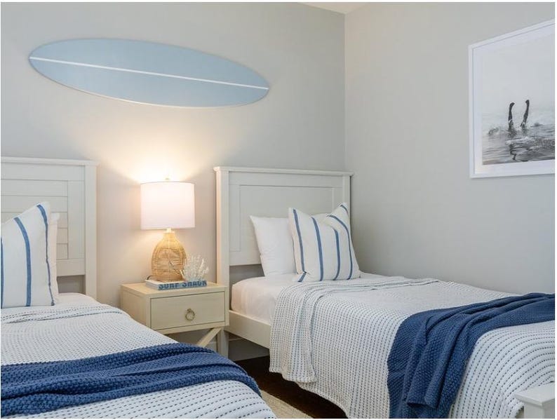 K&ouml;nnte beinhalten: Ein Schlafzimmer mit zwei wei&szlig;en Betten, blau-wei&szlig; gestreiften Kissen und marineblauen &Uuml;berw&uuml;rfen. &Uuml;ber den Betten h&auml;ngt eine hellblaue Surfbrett-Wanddekoration. Zwischen den Betten steht ein Nachttisch mit Lampe, an der Wand h&auml;ngt ein gerahmtes Bild.