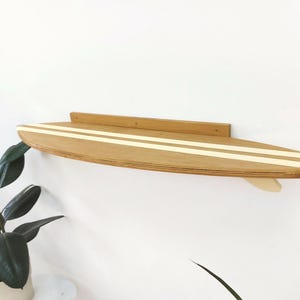 May include: A wooden surfboard shelf with two white stripes painted on the top. The shelf is mounted to a white wall.