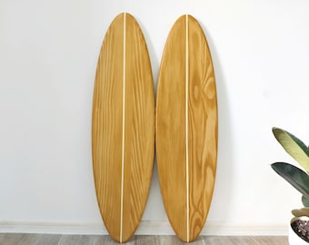 Op de afbeelding: Twee houten surfplank wanddecoraties in een lichtbruine kleur met een witte streep in het midden. De surfplanken hangen aan een witte muur.