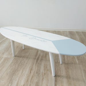 May include: White and light blue surfboard-shaped coffee table with four white legs. The table has a light blue section on one end and the text "the Bluewater" in the centre. The table is approximately 150 cm long.