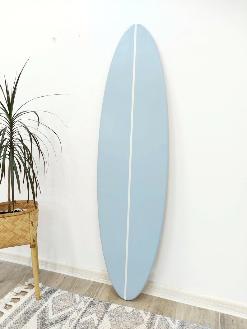 K&ouml;nnte beinhalten: Ein hellblaues Surfbrett mit einem einzelnen vertikalen wei&szlig;en Streifen in der Mitte. Das Brett hat eine glatte, matte Oberfl&auml;che sowie eine abgerundete Nase und ein abgerundetes Heck, was ein schlichtes Design mit Fokus auf die Farbe ergibt.