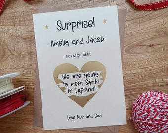May include: A white card with gold lettering that reads "Surprise! Amelia and Jacob" and "We are going to meet Santa in Lapland!" with a gold heart shape with a scratch-off surface. The card is on a brown surface with red and white string.