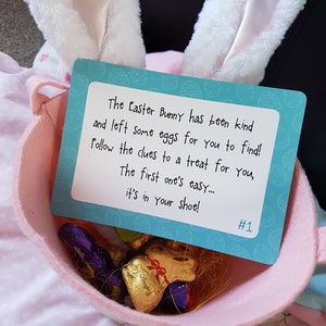 May include: A pink Easter basket with a blue card that reads "The Easter Bunny has been kind and left some eggs for you to find! Follow the clues to a treat for you, The first one's easy... it's in your shoe! #1". The basket contains chocolate bunny-shaped candies.