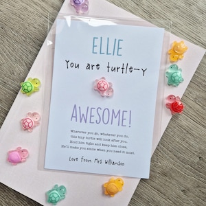 May include: A white card with the text "ELLIE You are turtle-y AWESOME!" and a poem, featuring a small pink turtle figurine attached to the centre. The card is surrounded by several colourful, translucent miniature turtle figurines in various colours.