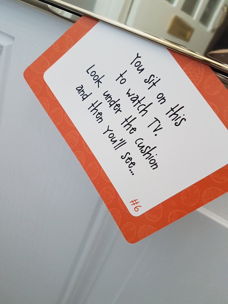 May include: A white card with orange borders and a pattern of eggs. The card has the text "You sit on this to watch TV. Look under the cushion and then you'll see... #6" written on it.