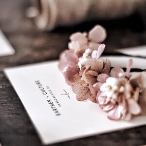 May include: Two hair clips with delicate pink dried flowers attached to black bobby pins. The flowers are arranged in a cluster and have a soft, romantic look. The card in the background reads "Earthen + Culture Handcrafted by..."