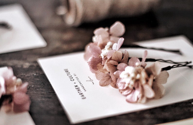 May include: Two hair clips with delicate pink dried flowers attached to black bobby pins. The flowers are arranged in a cluster and have a soft, romantic look. The card in the background reads "Earthen + Culture Handcrafted by..."