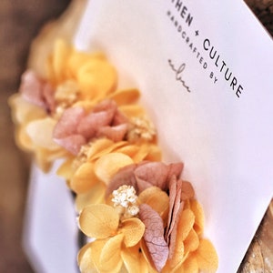May include: A hair clip with dried flowers in shades of yellow and pink. The flowers are arranged in a cluster and are attached to a black metal clip. The clip is labeled "Earthen + Culture Handcrafted by"