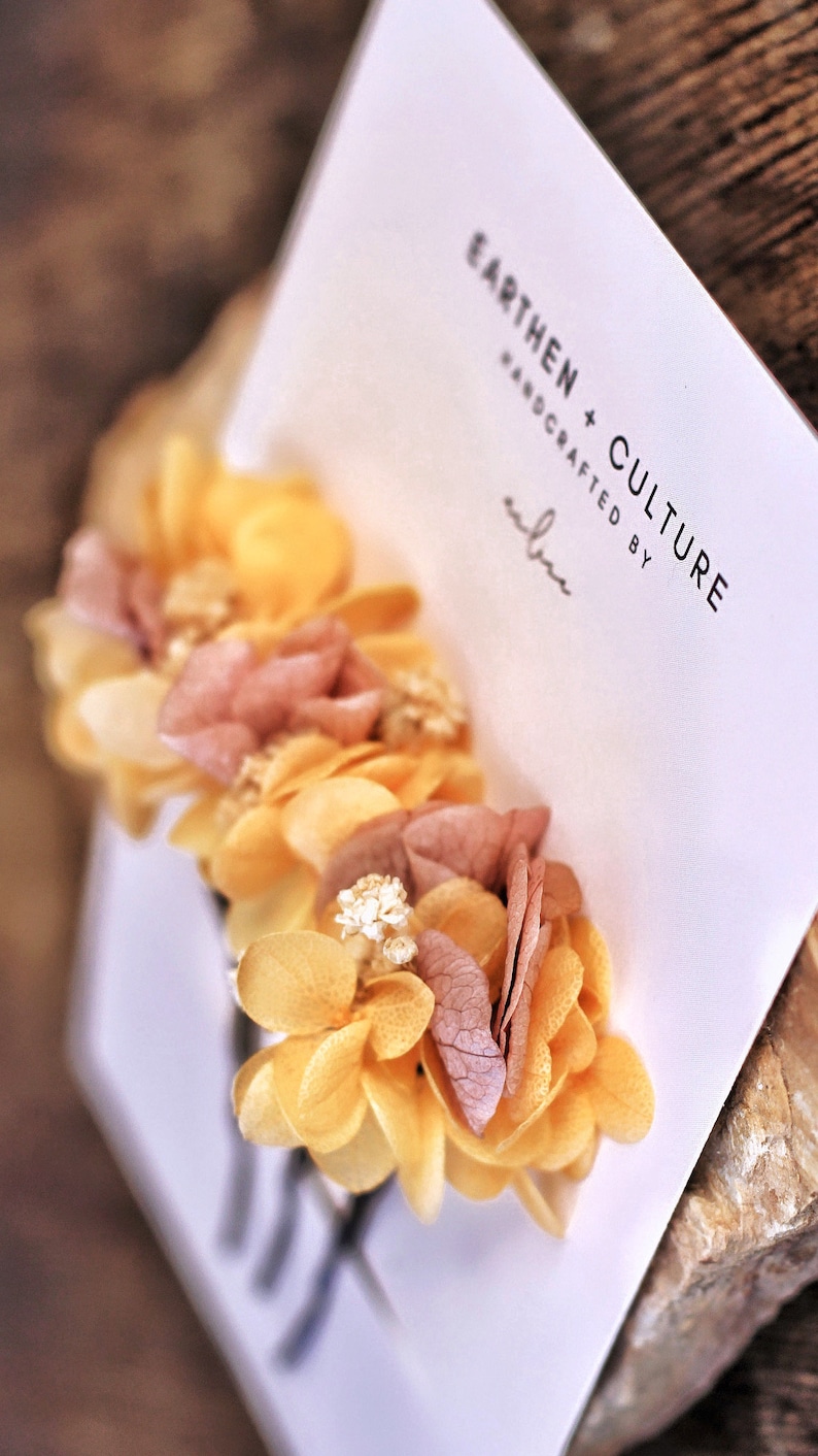 May include: A hair clip with dried flowers in shades of yellow and pink. The flowers are arranged in a cluster and are attached to a black metal clip. The clip is labeled "Earthen + Culture Handcrafted by"