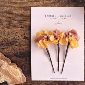 May include: Three hair clips with dried flower accents. The flowers are a mix of yellow and pink. The card reads "Earthen + Culture Handcrafted by... www.earthenandculture.com"