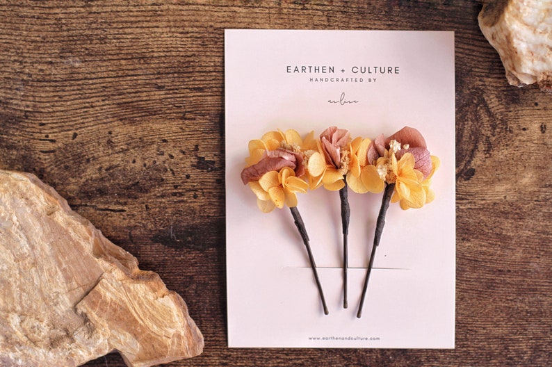 May include: Three hair clips with dried flower accents. The flowers are a mix of yellow and pink. The card reads "Earthen + Culture Handcrafted by... www.earthenandculture.com"