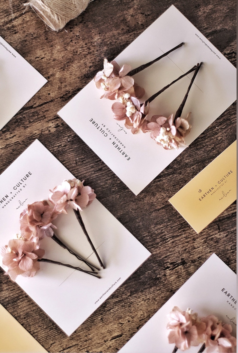 May include: Three sets of hair clips with dried pink flowers and a white card with the text "EARTHEN CULTURE HANDCRAFTED BY arlin" and the website address "www.earthasandculture.com".