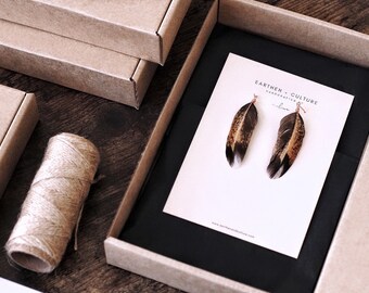 May include: A pair of feather earrings on a white card with the text "Earthen Culture Handcrafted" and a website address. The earrings are made of brown and white feathers with gold accents.