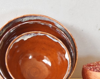May include: Three brown ceramic bowls of varying sizes are stacked on top of each other on a wooden surface. A half of a pomegranate with red seeds is next to the bowls.