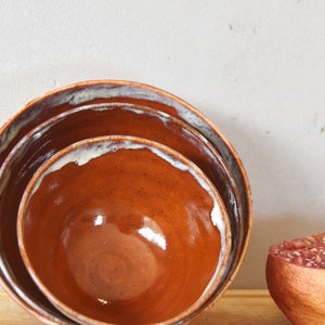 Peut inclure: Trois bols en céramique brune de tailles différentes sont empilés les uns sur les autres sur une surface en bois. Une demi-grenade aux graines rouges se trouve à côté des bols.