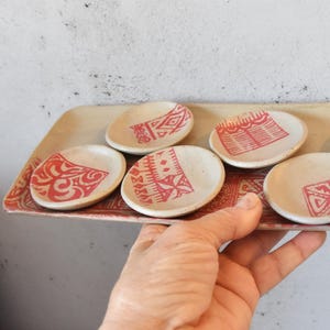 Könnte beinhalten: Ein Set aus sechs kleinen, weißen Keramiktellern mit roten geometrischen Mustern. Die Teller sind auf einem größeren, rechteckigen, weißen Keramiktablett mit einem roten geometrischen Muster angeordnet.