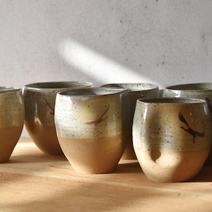 May include: A set of handmade ceramic tumblers. Each tumbler has a speckled, light-coloured upper section and a tan lower section. A dark brown bird silhouette is painted on each tumbler. The tumblers are arranged on a wooden surface.