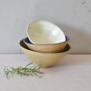 Pode incluir: Três tigelas de cerâmica de tamanhos variados são empilhadas. A tigela maior é bege salpicado, a do meio tem uma borda azul e a menor é branca. Um raminho de alecrim está em primeiro plano.