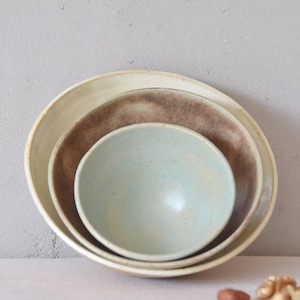 Pode incluir: Um conjunto de três tigelas de cerâmica de tamanhos diferentes, encaixadas umas nas outras. A tigela maior é branco-sujo, a do meio é castanha e a menor é azul claro. As tigelas são provavelmente para servir alimentos.