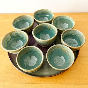 May include: Eight small ceramic bowls with a teal interior and a speckled, light brown exterior are arranged on a dark purple and green plate. The bowls are handmade and have a rustic, artisanal appearance.