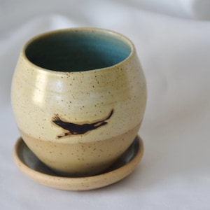 Puede incluir: Una taza de cerámica beige moteada con interior verde azulado oscuro y platillo a juego. Una silueta de pájaro negro está pintada en la taza. La taza tiene forma redondeada.