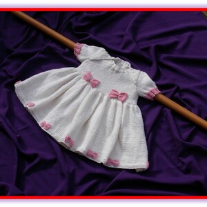 May include: A white knitted baby dress with short sleeves and a flared skirt. The dress is adorned with pink bows and trim. The dress is displayed on a purple fabric background with a wooden dowel.