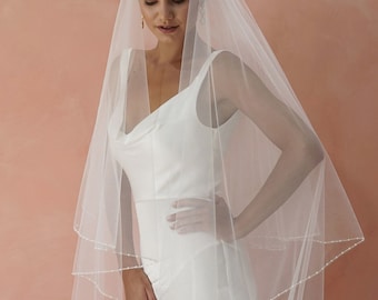 May include: A long, sheer white bridal veil featuring a delicate trim of small, evenly spaced white beads along the edges. The veil is draped over a sleeveless white gown with a cowl neckline and a high front slit, creating a soft, layered look.