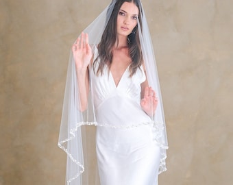 May include: A floor-length white satin slip dress with a deep V-neckline paired with a sheer white tulle veil. The veil features a delicate scalloped edge adorned with small white beads that follow the perimeter of the fabric in a continuous pattern.