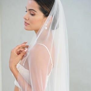 May include: A bride wearing a white wedding dress and veil. The sheer veil drapes over the shoulders and back. The bride is wearing pearl earrings and a matching white top and skirt. The image is taken in a studio setting with soft lighting.
