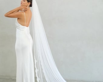 May include: A floor-length white satin slip dress with thin spaghetti straps and a low back, paired with a long, sheer white bridal veil featuring a delicate scalloped lace trim along the edges that trails onto the floor behind the garment.