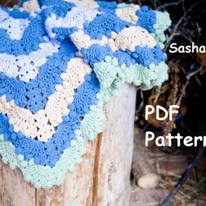 Puede incluir: Una manta de bebé de ganchillo en tonos de azul, crema y verde. La manta está hecha con un punto texturizado y tiene un borde festoneado. La manta está drapeada sobre un tronco de madera. El texto "SashaPatern PDF Pattern" es visible en la imagen.