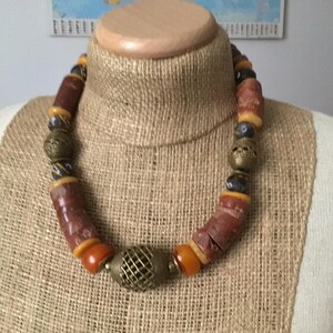 May include: A statement necklace with a rustic aesthetic. It features large, cylindrical brown beads, complemented by orange and yellow beads, and brass-toned metal beads. A prominent cage-like bead serves as the central focus of the necklace.