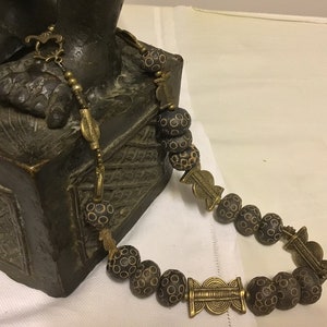 Può includere: Una collana artigianale con perline marrone scuro decorate e dettagli in metallo dorato. La collana è drappeggiata su una scultura in bronzo, evidenziando il suo design intricato. La lunghezza della collana è di circa 50 cm.