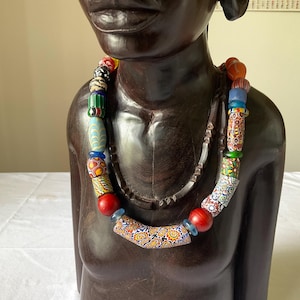 Puede incluir: Un collar de cuentas de colores con una variedad de formas y tamaños. El collar presenta cuentas rojas, azules, verdes y estampadas. El collar se exhibe en un busto de madera marrón oscuro.
