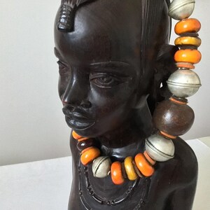 Può includere: Una scultura in legno marrone scuro della testa e della parte superiore del busto di una persona. La scultura ha un volto dettagliato ed è adornata da una collana di perline arancioni, gialle e argentate. La scultura è un'opera d'arte decorativa.
