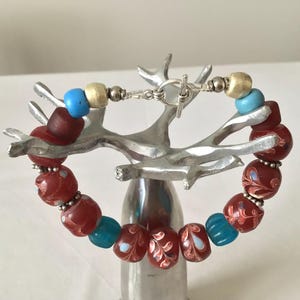 Può includere: Bracciale di perline con chiusura a toggle. Presenta perline rosse, blu e dorate. Le perline rosse hanno un motivo floreale. Il bracciale è esposto su un supporto decorativo argentato.