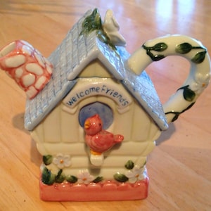 May include: A white ceramic teapot shaped like a birdhouse with a blue roof, pink trim, and a green vine handle. The teapot has a red bird inside and the words "Welcome Friends" written on the front.