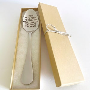 K&ouml;nnte beinhalten: Ein silberner L&ouml;ffel mit der Gravur "We've been trying to reach you about your cars extended warranty". Der L&ouml;ffel befindet sich in einer braunen Geschenkbox mit einem wei&szlig;en Band.