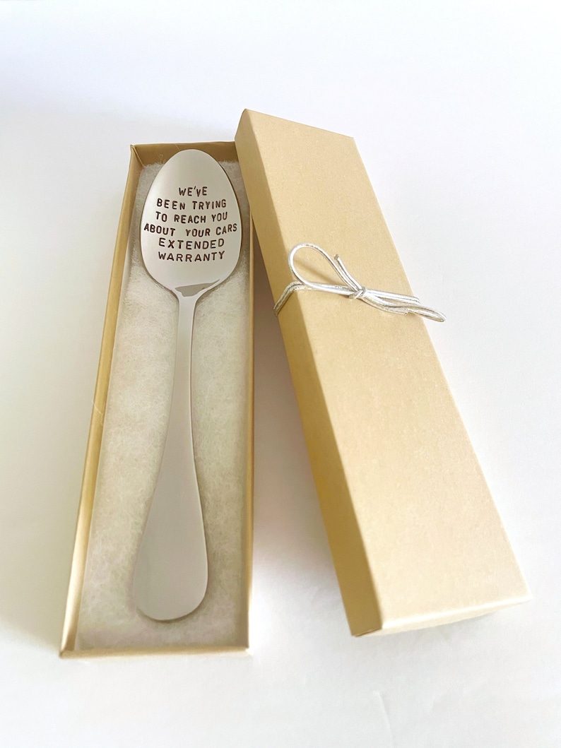 K&ouml;nnte beinhalten: Ein silberner L&ouml;ffel mit der Gravur "We've been trying to reach you about your cars extended warranty". Der L&ouml;ffel befindet sich in einer braunen Geschenkbox mit einem wei&szlig;en Band.
