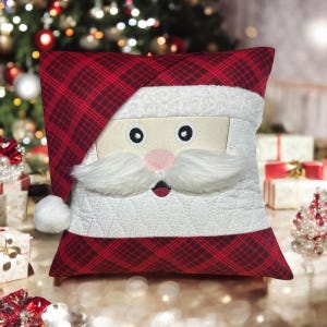 May include: A decorative Christmas pillow featuring Santa Claus's face. The pillow has a red plaid hat and base, with a white face, mustache, and beard. The eyes are black circles, and the nose is pink. The pillow is square-shaped.