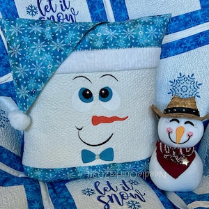 May include: A white snowman pillow with a blue hat and a blue bow tie. The pillow is sitting on a blue and white quilt with the words "Let it snow" embroidered on it. There is a small snowman figurine with a cowboy hat and red bandana sitting next to the pillow.