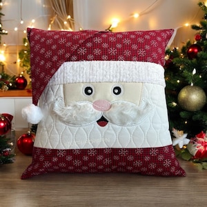 Peut inclure: Un coussin de Noël rouge et blanc avec un visage de Père Noël brodé sur le devant. Le coussin a un pompon blanc sur le dessus du chapeau.