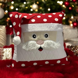 May include: Red and white polka dot pillow with a white Santa Claus face embroidered on it. The Santa Claus has a white beard and mustache, and a pink nose.